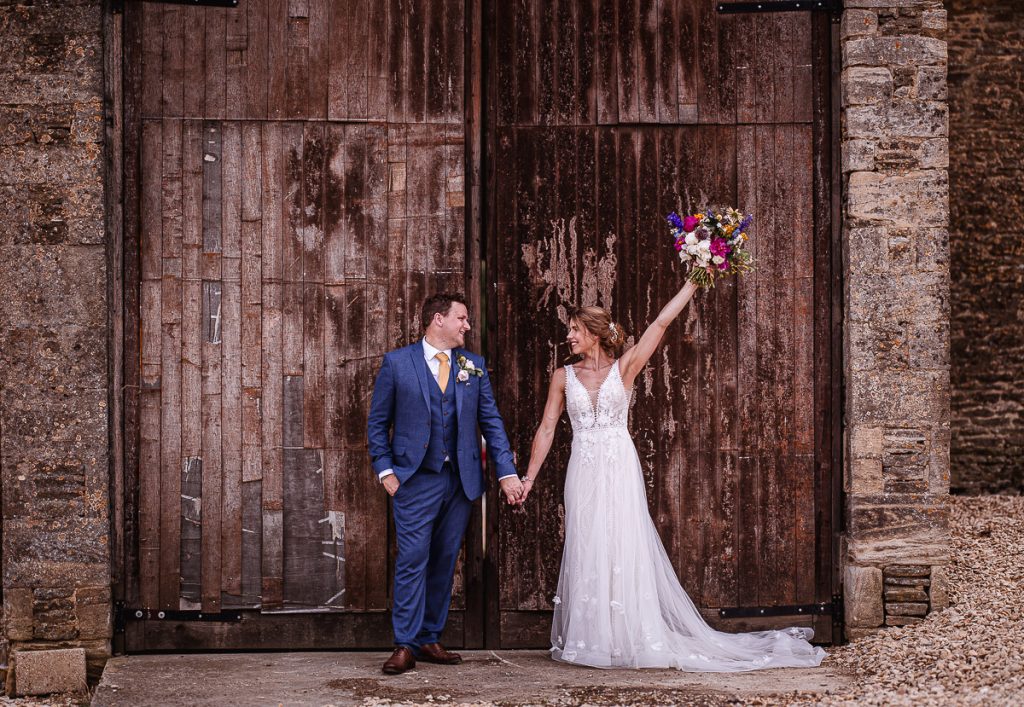 Photo of bride and groom standing infront of wooden rustic gate holing their hands and looking at each other