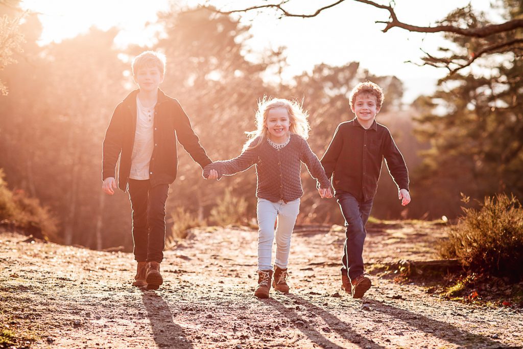 Three children holding hands and smiling as they walk outdoors on a sunlit, wooded path—a heartwarming moment captured in the signature style of Hampshire Wedding Photography, with sunlight streaming through the trees in the background.