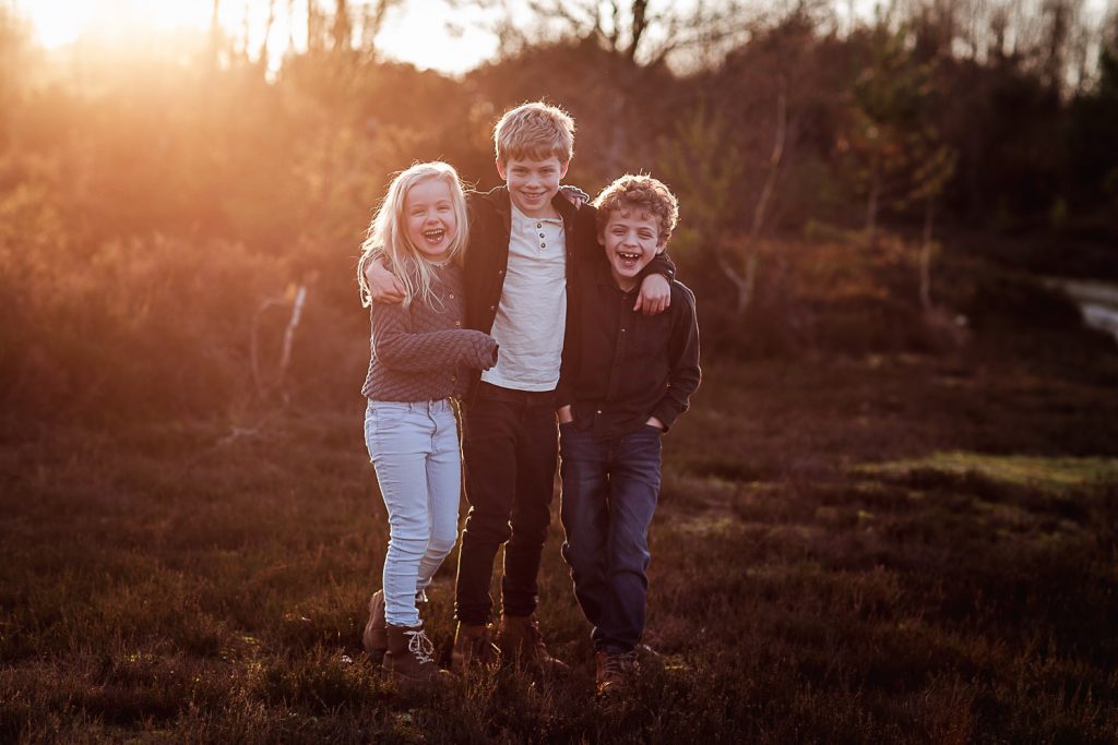 Three children stand outdoors at sunset, smiling and hugging each other, with trees and grass in the background. The warm, glowing atmosphere captures a joyful moment reminiscent of Hampshire Wedding Photography style.