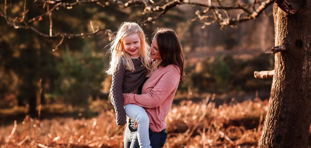 A woman in a pink sweater smiles while holding a young girl with blonde hair outdoors, surrounded by trees and brown autumn foliage—capturing a joyful moment in a style reminiscent of Hampshire Wedding Photography.