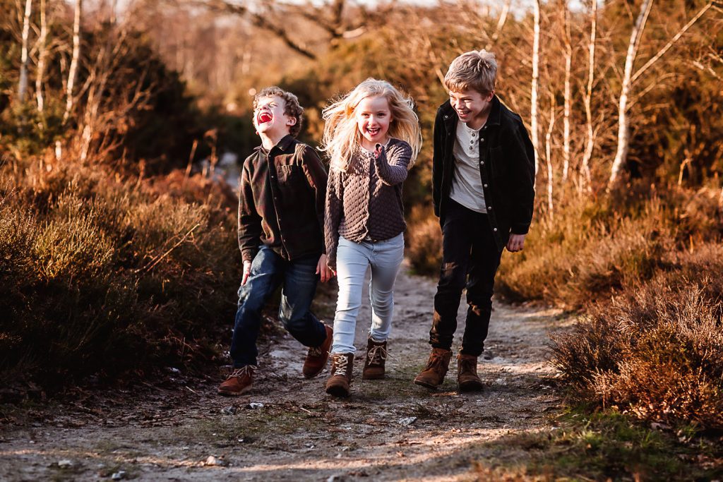 Three children walk and laugh together on a dirt path, surrounded by bushes and trees, enjoying a sunny day outdoors—a moment reminiscent of candid Hampshire Wedding Photography.