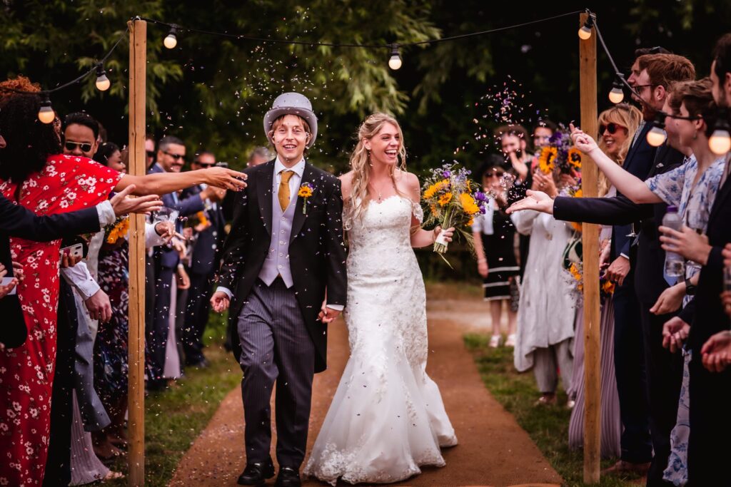 A bride and groom walk hand-in-hand down an outdoor aisle, smiling as guests shower them with confetti. String lights twinkle above, capturing the festive joy perfectly—an unforgettable moment for any Hampshire wedding photographer.