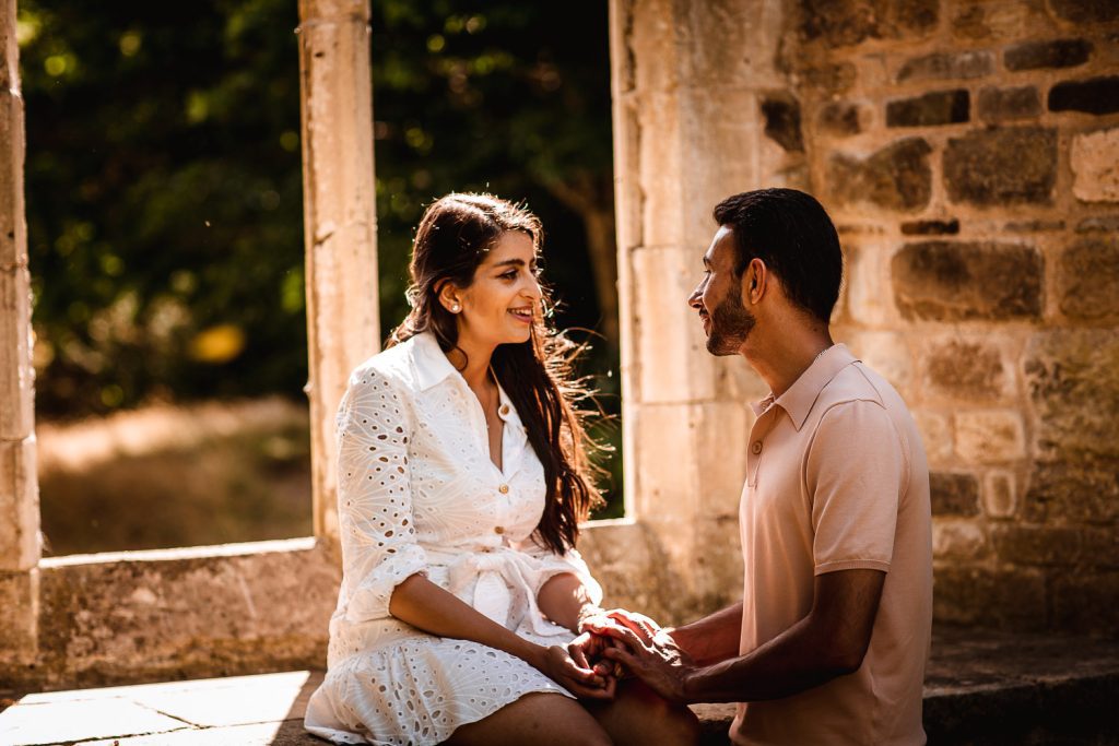 A couple sits together by a stone window, holding hands and smiling warmly. Sunlight streams in, creating a soft, romantic atmosphere—perfect inspiration for full day wedding packages that capture every cherished moment.