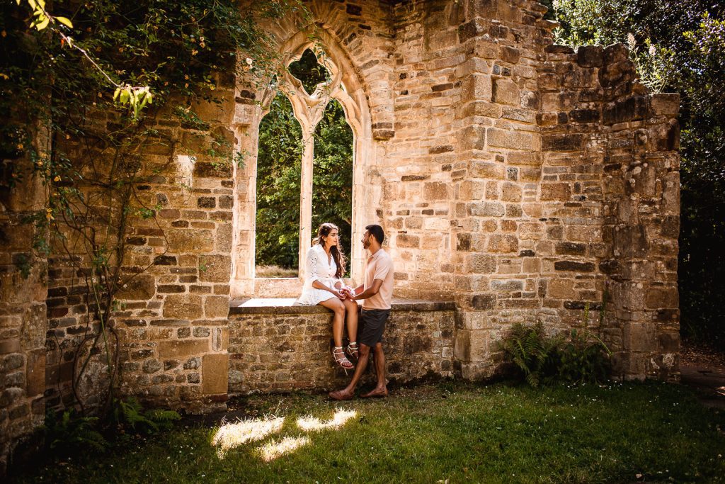 A couple sits and stands by a stone window of a historic, weathered building, holding hands and looking at each other—captured in golden sunlight amidst greenery. Discover full day wedding packages to relive moments like these forever.