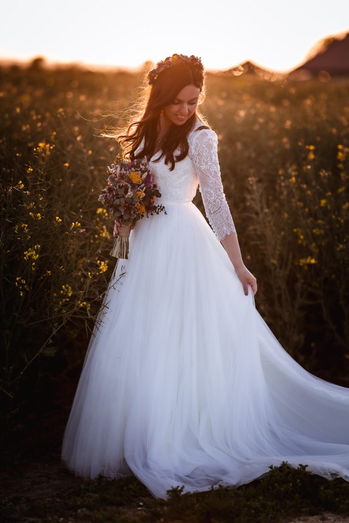 sunset photo of bride holding her flowers and looking down at her dress in the field at Chidham Barn, Chichester wedding photographer