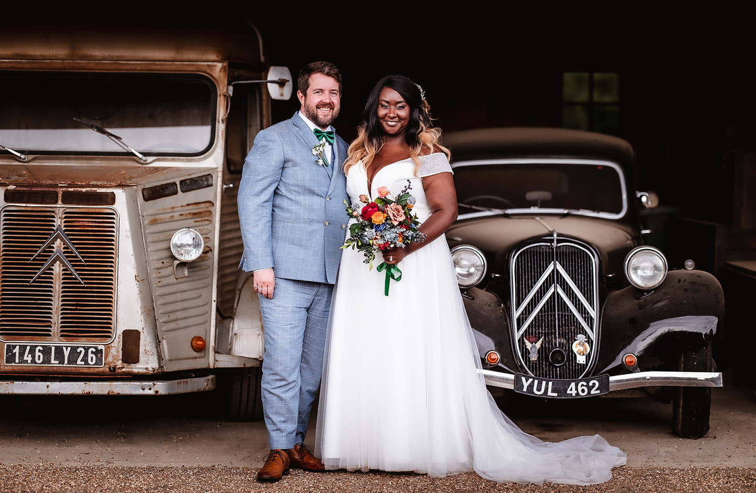 Bride and groom standing behind old cars at the barn