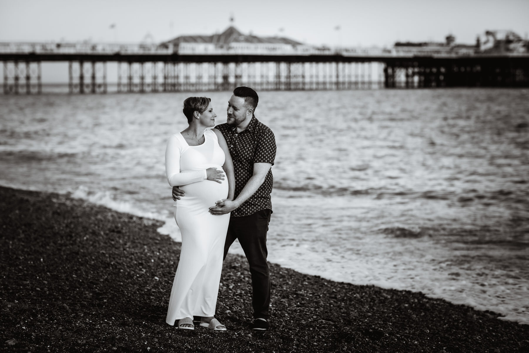 A couple stands on a pebbled beach, with a pier in the background. The pregnant woman in a white dress holds her belly, while her partner embraces her, both looking at each other and smiling. The scene is in black and white.