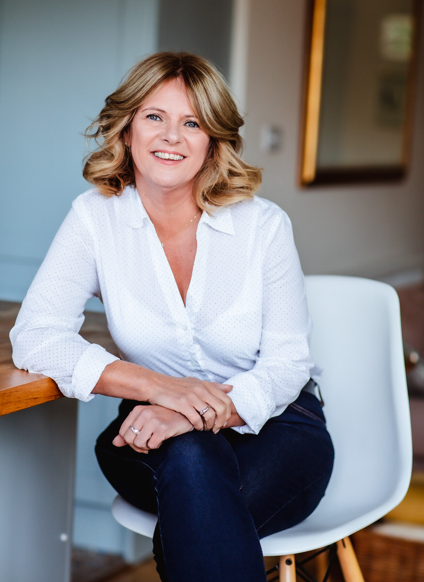 A woman with blonde, shoulder-length hair sits on a white chair, smiling. She wears a white button-up shirt and dark jeans, with her arms resting casually on a table in a bright, modern room.