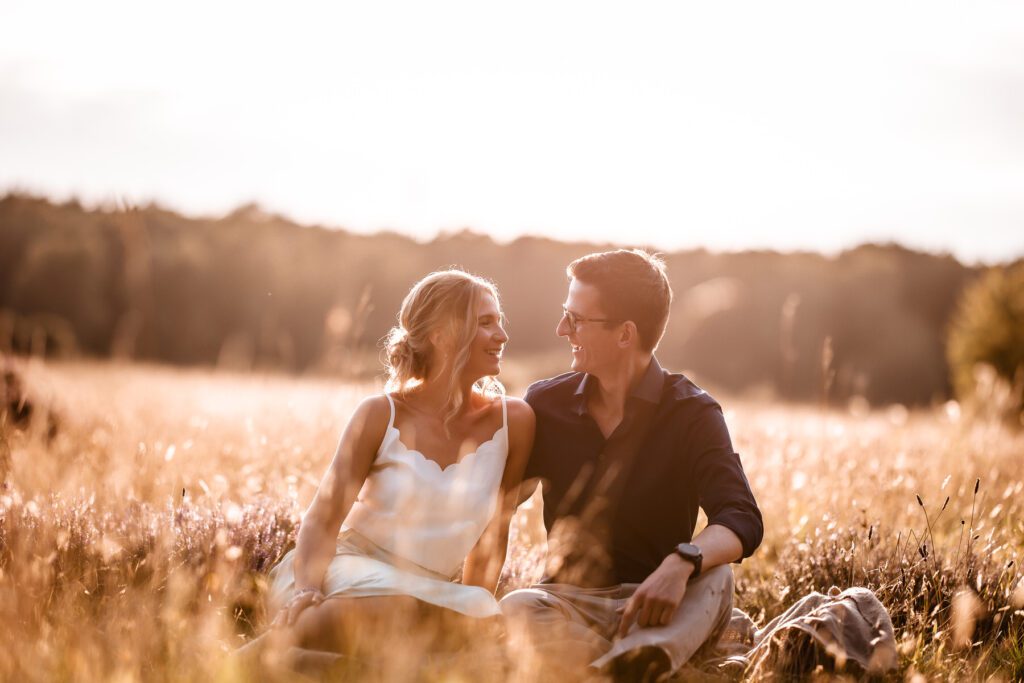 A couple sits close together in a sunlit field, smiling at each other. The woman wears a white dress and the man wears a dark shirt. The scene is warm and romantic, with tall grass and trees in the background.