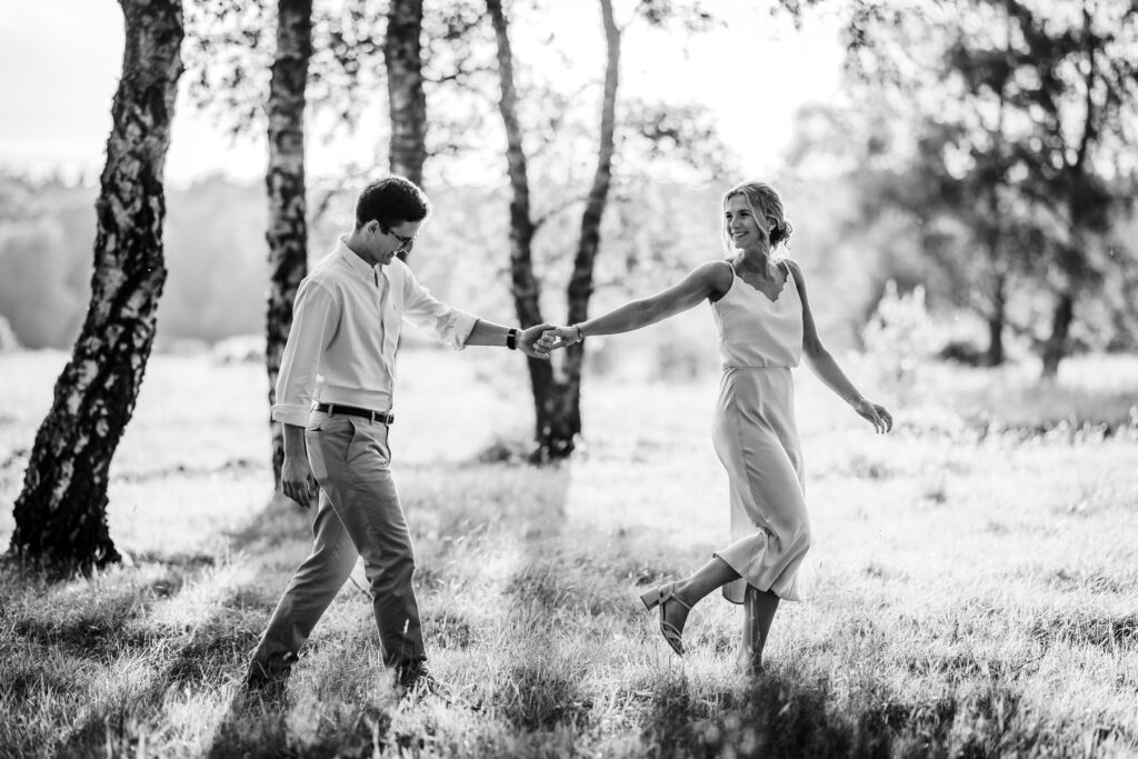 A couple walks hand in hand through a sunlit grassy field, smiling and looking at each other. Tall trees and soft sunlight create a peaceful, romantic atmosphere. The image is in black and white.