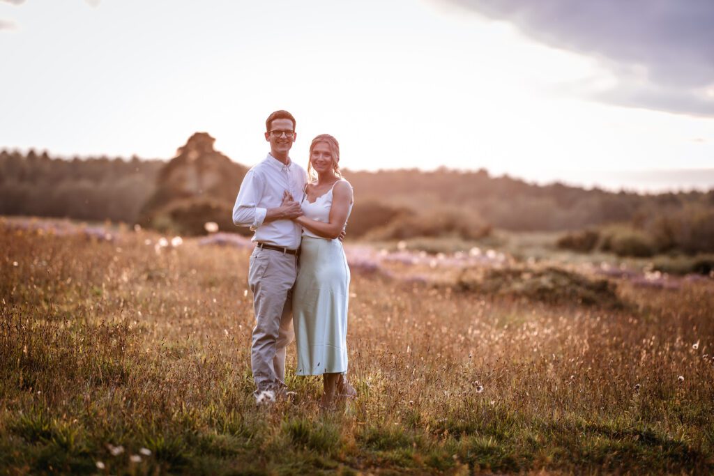 A smiling couple stands closely together in a sunlit field with tall grass and wildflowers, with trees and a soft, glowing sky in the background.