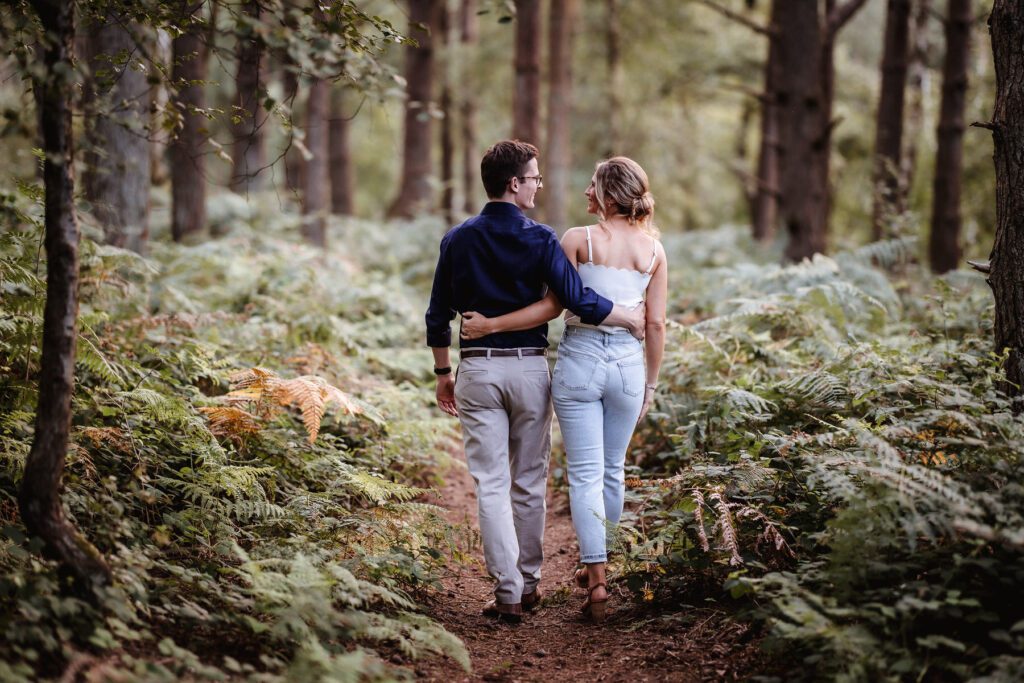 A couple is walking arm-in-arm down a narrow dirt path surrounded by lush green ferns and tall trees in a forest. They are viewed from behind and appear relaxed and close.
