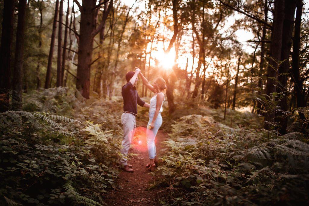 A couple dances on a forest path surrounded by lush greenery, with sunlight streaming through the trees and casting a warm glow around them.