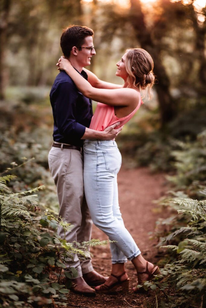 A couple stands closely together on a forest path, embracing and smiling at each other. Sunlight filters through the trees, creating a warm, romantic atmosphere. The woman wears a pink top and jeans; the man wears a dark shirt and khakis.