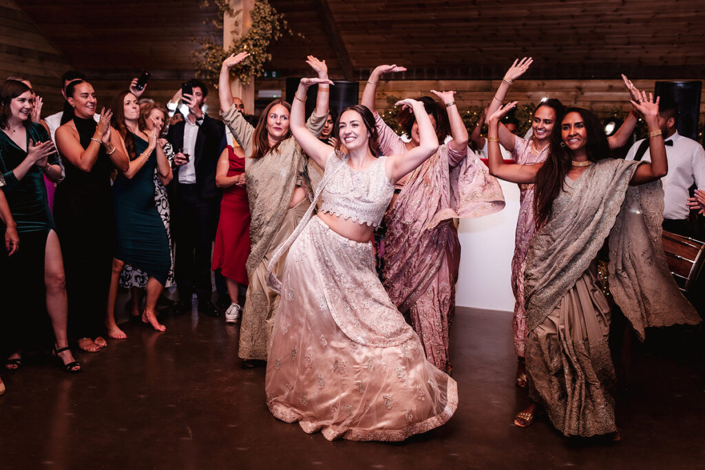 bride is dancing indian dance with her bridesmaids at indoor rusting wedding venue in Surrey called High Billinghurst Farm