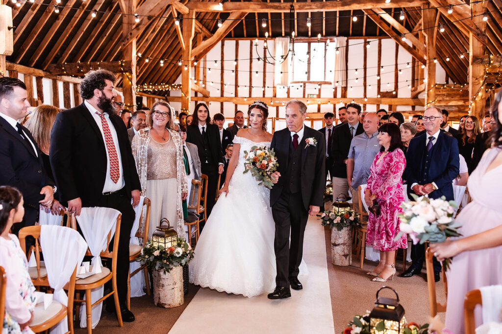 bride is coming down the isle with her dad at rustic Clock Barn Hampshire wedding venue