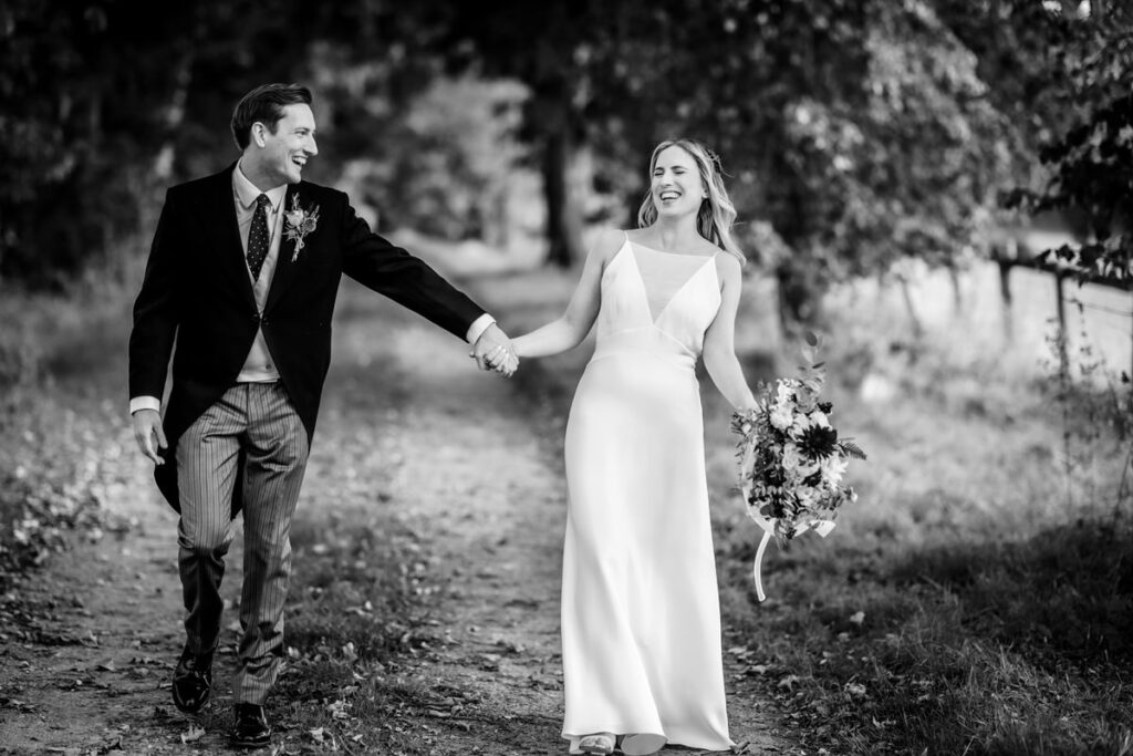 black and white photo of bride and groom walking together on the pathway within the trees having fun and laugh 