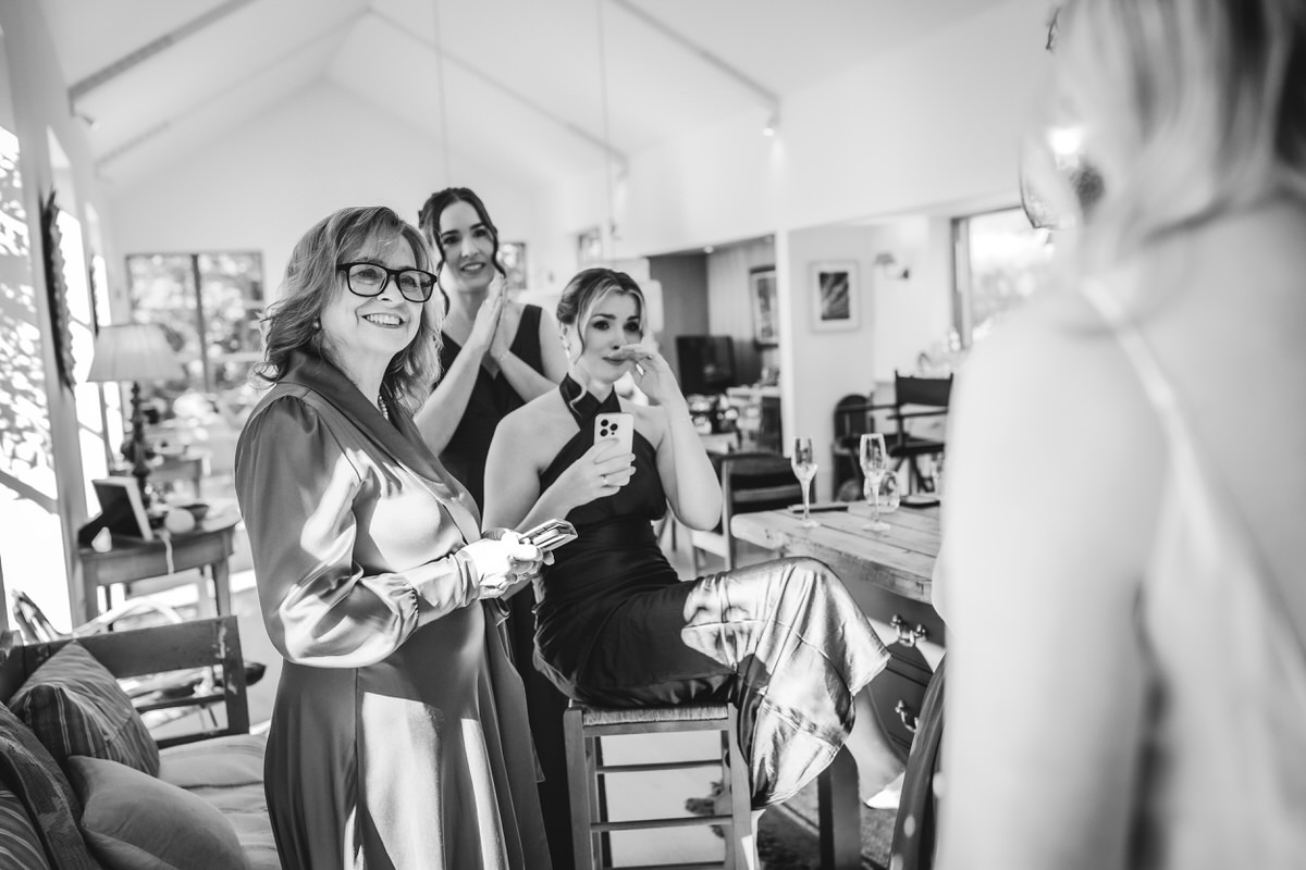 A group of women in formal attire gather in a bright, modern room. One woman smiles at the camera while others engage in conversation, holding drinks. The atmosphere appears joyful and celebratory.