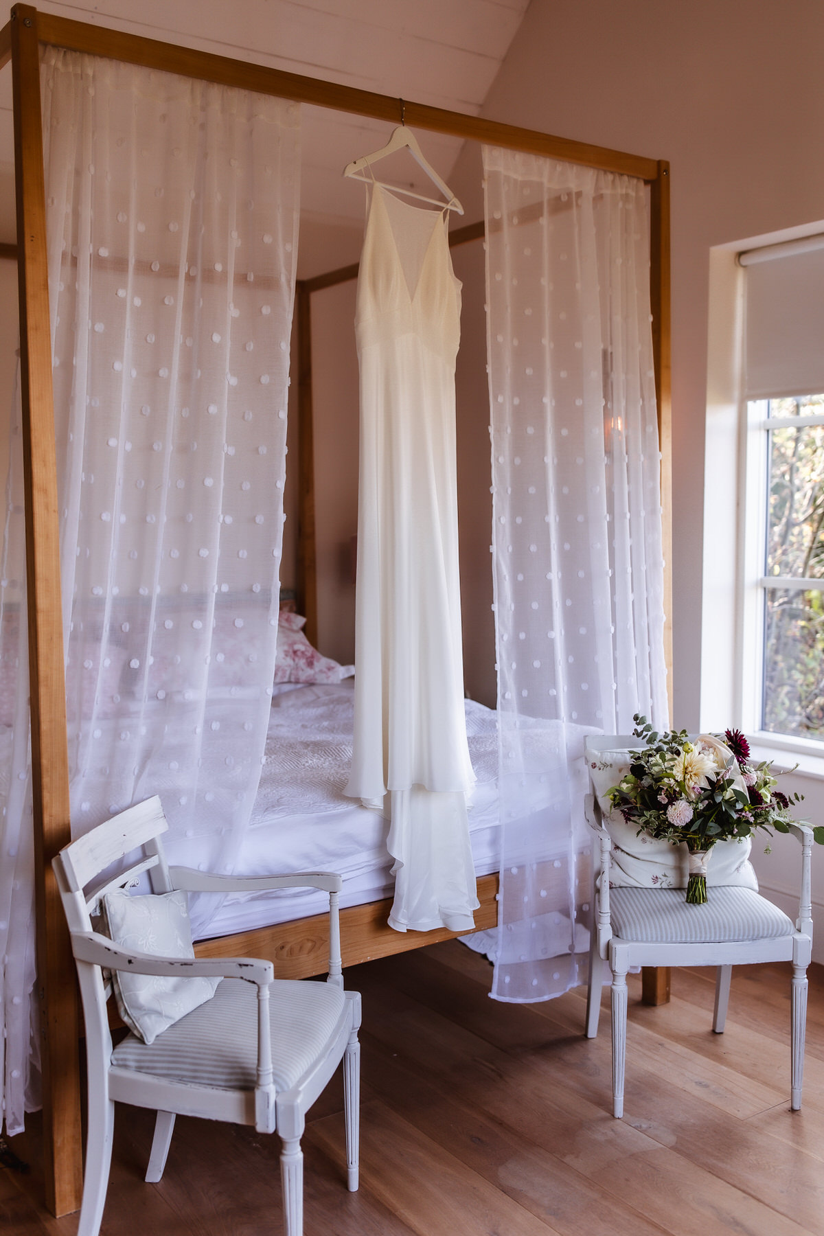 A long white wedding dress hangs from a four-poster bed draped with sheer, polka-dot curtains. Two white chairs sit nearby, one holding a bouquet of white and burgundy flowers. Sunlight enters through a window.