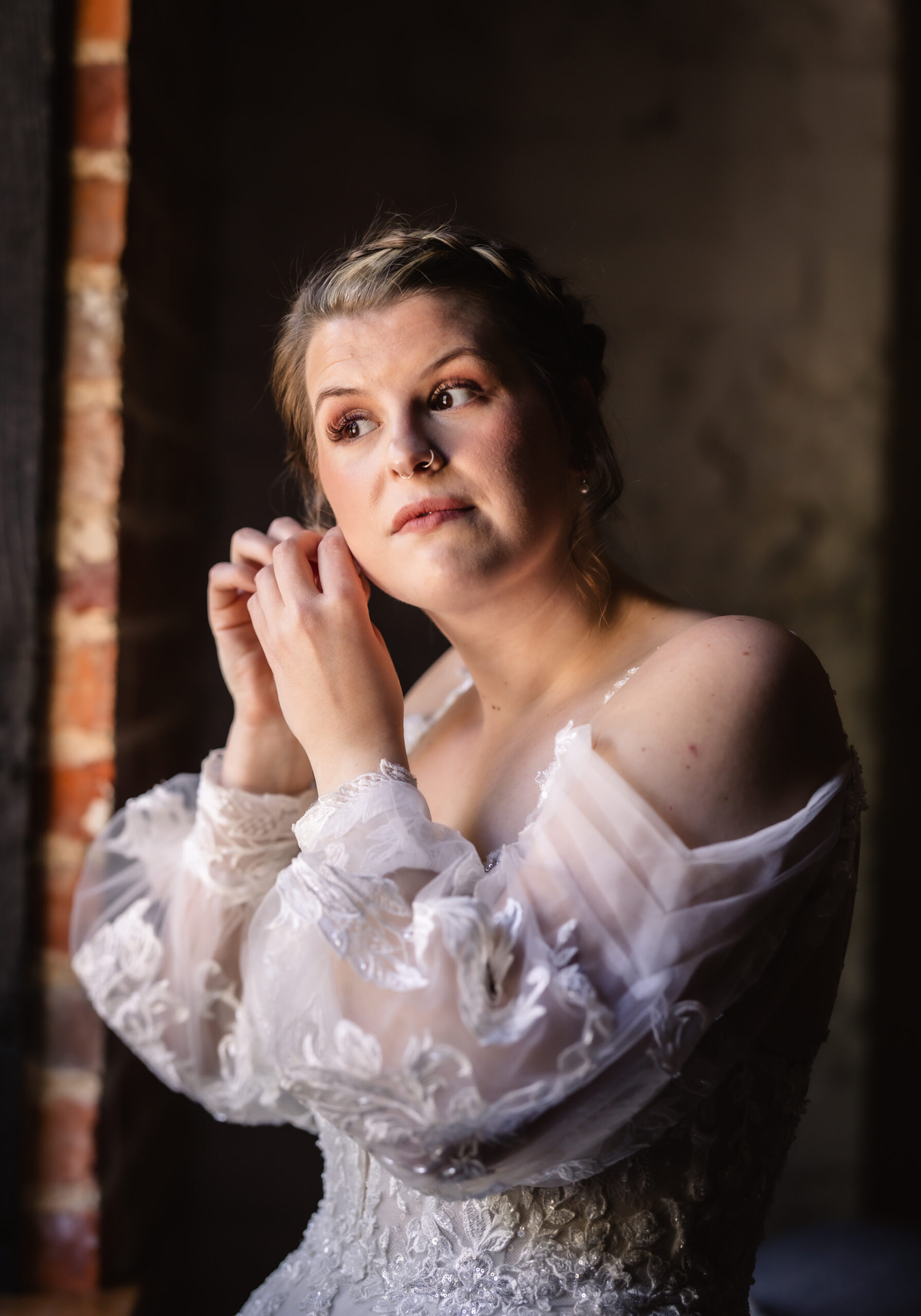 The bride stands by the window, where soft morning light filters gently into the room, illuminating the space in a warm glow. She takes a deep breath, feeling a mix of excitement and nervousness as she reaches for her earrings. The delicate jewels sparkle in the light, catching her eye as she holds them close.