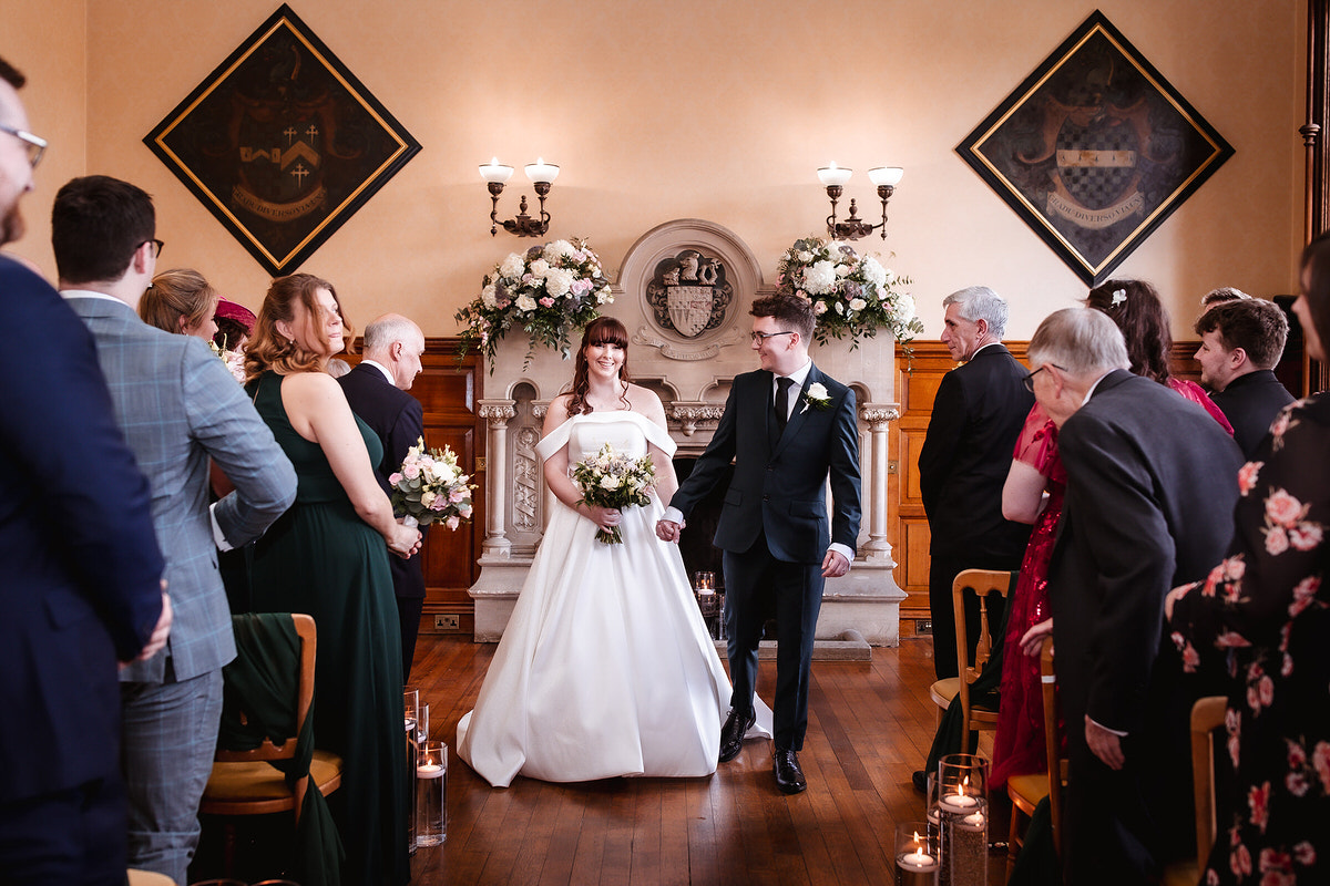 A bride and groom hold hands and smile as they walk down the aisle in an elegant indoor venue, surrounded by seated guests. The room features floral arrangements and ornate wall decorations.