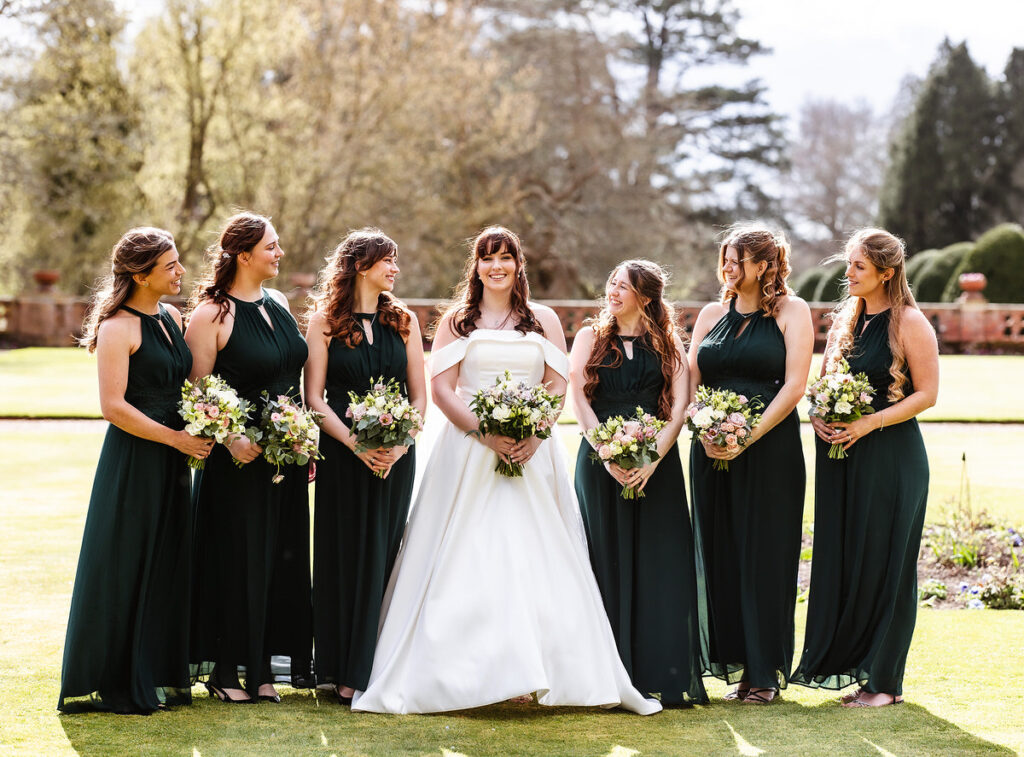 A bride in a white gown stands outdoors with six bridesmaids in matching dark green dresses, each holding a bouquet. They are smiling and standing on grass, with trees and sunlight in the background.