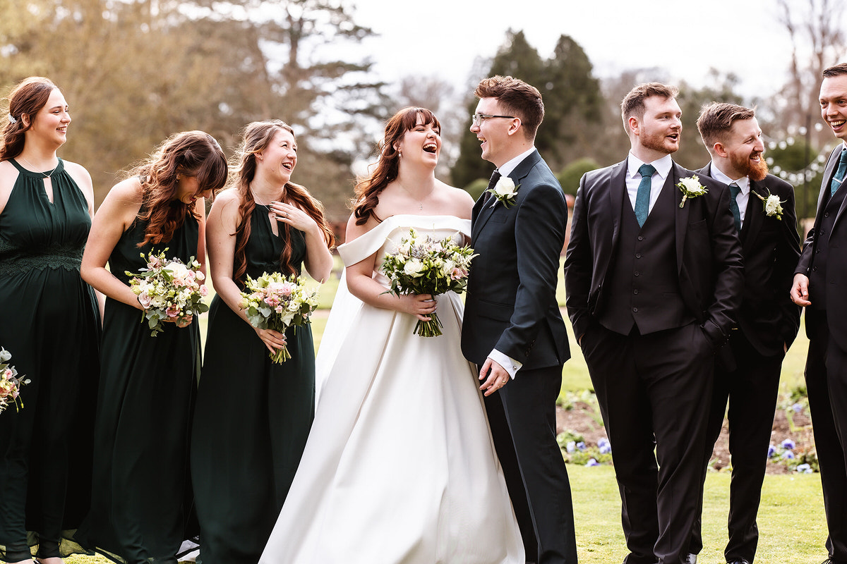 A bride and groom stand outdoors, smiling and laughing with their bridesmaids and groomsmen, all dressed in formal wear. The group holds bouquets, and trees are visible in the background.