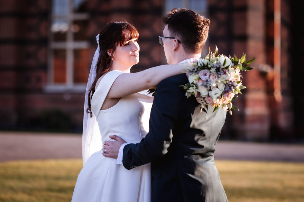 A bride in a white dress and veil embraces a groom in a dark suit outdoors, holding a bouquet of flowers. They gaze at each other with soft sunlight and a brick building in the background.