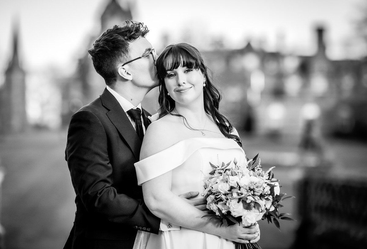 A groom in a suit kisses the bride’s temple as she smiles, holding a bouquet of flowers. They stand close together outdoors, dressed in wedding attire, with a blurred building in the background.