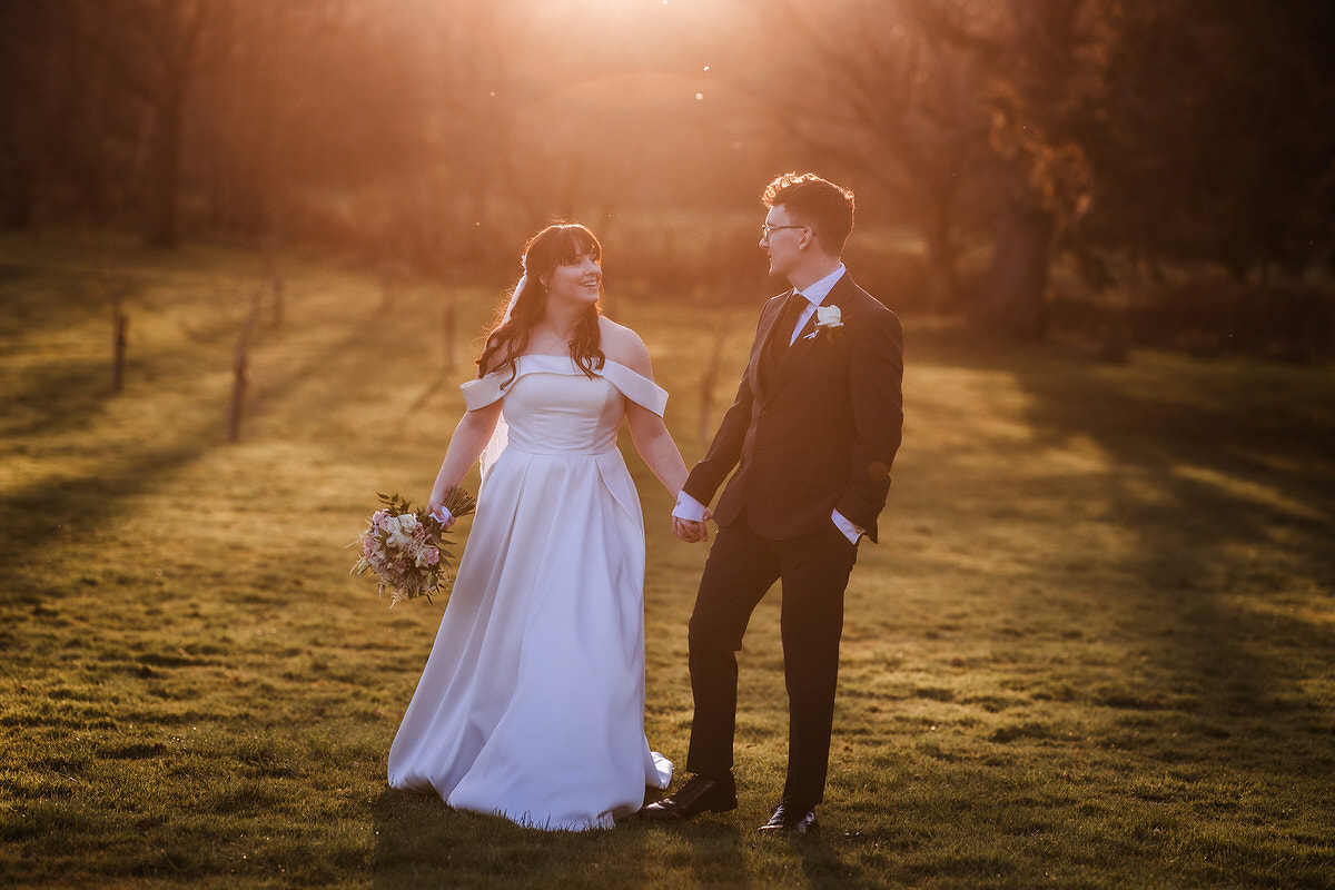 A bride in a white dress holding a bouquet and a groom in a suit hold hands and smile at each other while standing on grassy ground at sunset. Warm sunlight creates a romantic, golden glow around them.