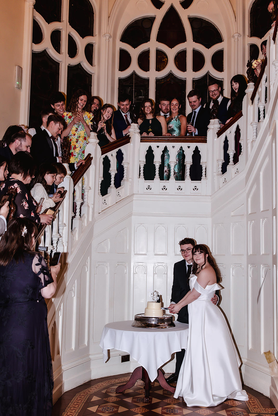 A bride and groom stand together, smiling, as they cut their wedding cake at the base of a grand staircase. Friends and family gather on the stairs, watching and taking photos in an elegant, historic venue.
