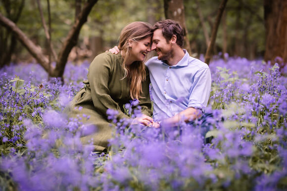 A couple sits closely together in a field of blooming purple flowers, smiling and touching foreheads, surrounded by trees in a peaceful, natural setting.