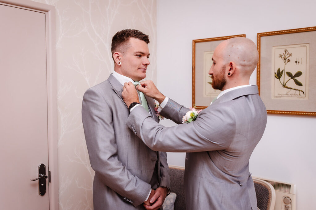 Two men in gray suits stand indoors. One adjusts the others tie, who stands with hands clasped, smiling. The room has beige walls, framed botanical art, and a closed door.