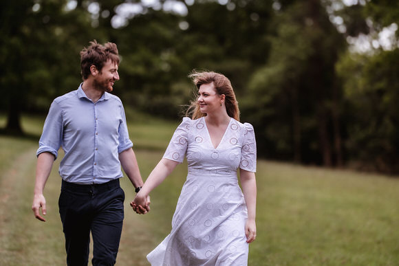 A man and woman holding hands walk through a grassy field, both smiling at each other. The woman wears a white dress and the man wears a light blue shirt and dark pants. Trees and greenery are in the background.