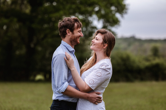 A couple stands outdoors, smiling at each other and embracing. The woman wears a white dress and the man wears a light blue shirt. They are surrounded by green grass and trees, with a cloudy sky in the background.