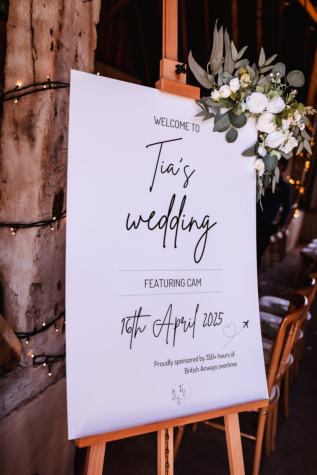 A white wedding welcome sign on an easel reads “Welcome to Tia’s wedding featuring Cam, 16th April 2025.” The sign has a small bouquet of white flowers and greenery at the top right corner. Wooden chairs and fairy lights are in the background.