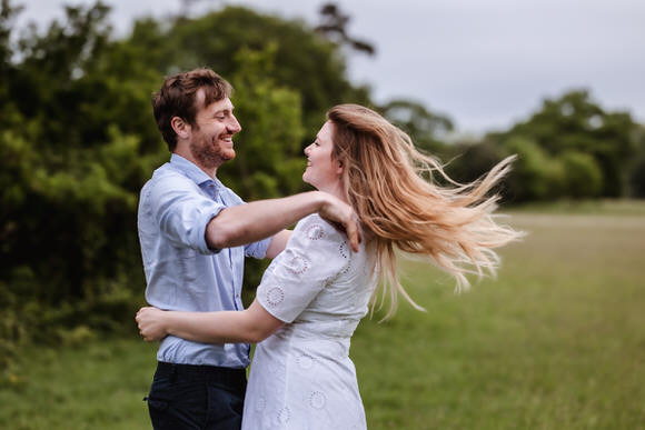 A smiling couple stands in a grassy field, embracing and looking at each other. The woman’s long hair flows in the wind, and both appear happy and relaxed, surrounded by green trees under a cloudy sky.
