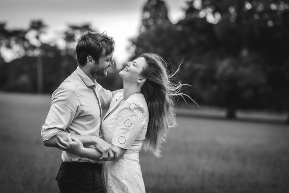 A smiling couple stands close together in a grassy field, laughing and holding each other. The woman wears a light dress and her hair blows in the wind. The photo is in black and white, with trees blurred in the background.