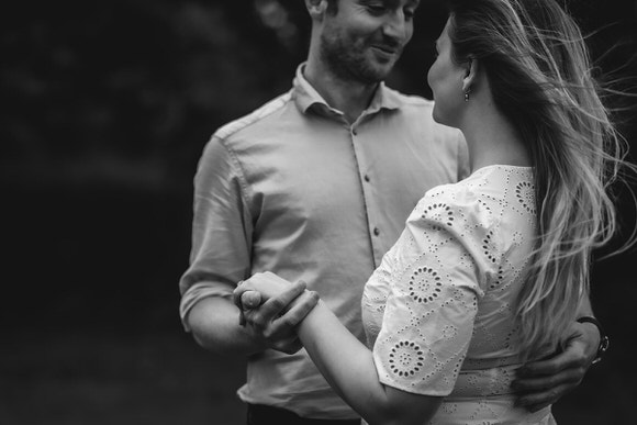 A black and white photo of a couple holding hands and smiling at each other. The woman’s hair is blowing in the wind, and they appear to be dancing outdoors.