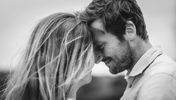 A man and woman stand close together, smiling with their foreheads touching. The image is in black and white, and they appear happy and affectionate.
