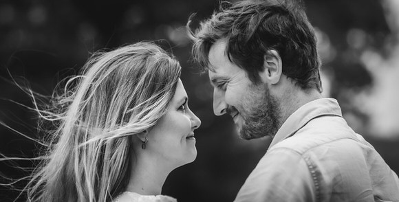 A black-and-white photo of a couple facing each other closely outdoors, smiling and gazing into each other’s eyes, with the womans hair blowing gently in the wind.