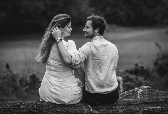 A couple sits close together on a log outdoors, facing each other and smiling lovingly. The woman wears a light dress, and the man gently touches her hair. The background is blurred with trees and grass.