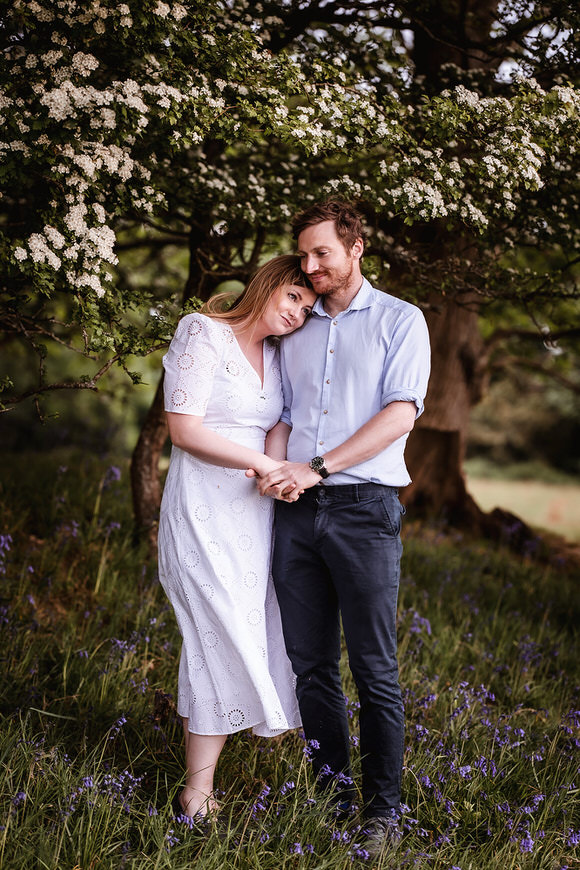 A couple stands close together under a tree with white blossoms. The woman in a white dress leans her head on the man’s shoulder, and he rests his head on hers. They hold hands and stand among grass and purple wildflowers.
