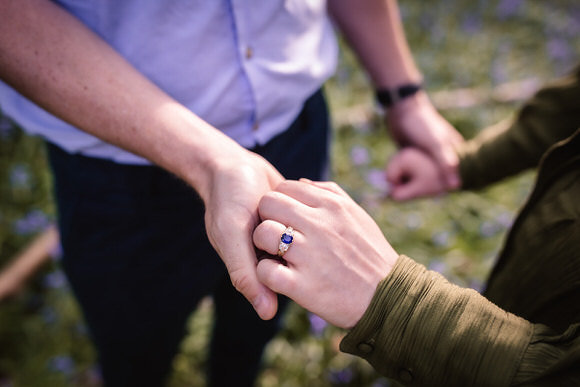 A close-up of two people holding hands outdoors, one wearing an engagement ring with a blue stone and a silver band, while the background is blurred with greenery and hints of purple flowers.