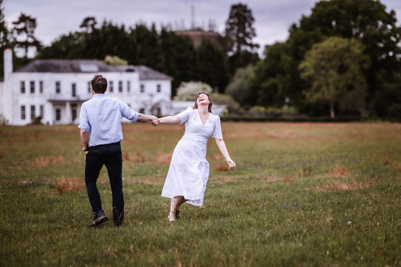 A man and woman holding hands and laughing as they run across a grassy field, with a large white house and trees in the background under a cloudy sky.