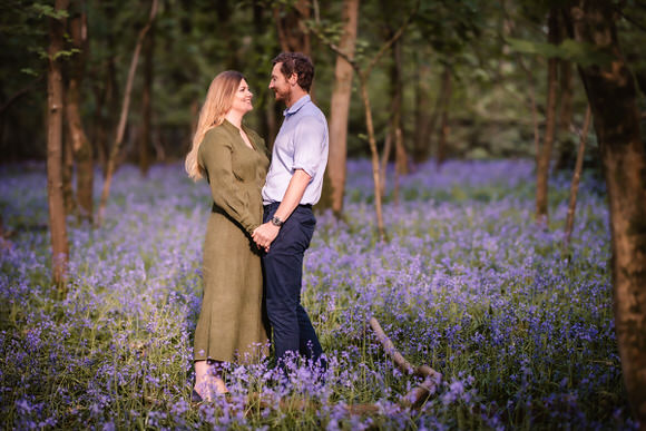 A couple stands holding hands and smiling at each other in a forest clearing filled with blooming purple flowers and surrounded by trees.