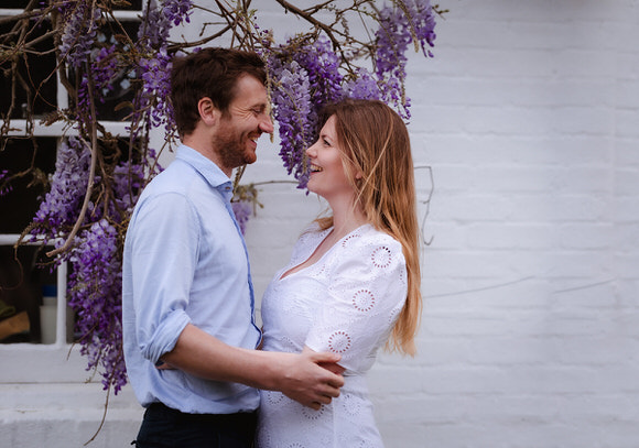A couple stands close together, smiling at each other under hanging purple wisteria flowers, in front of a white brick wall with a window. The man wears a light blue shirt and the woman wears a white dress.