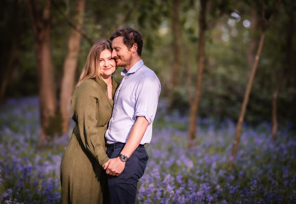 A couple stands close together, holding hands and smiling in a sunlit forest with purple wildflowers covering the ground. The woman wears a green dress, and the man wears a light blue shirt and dark pants.