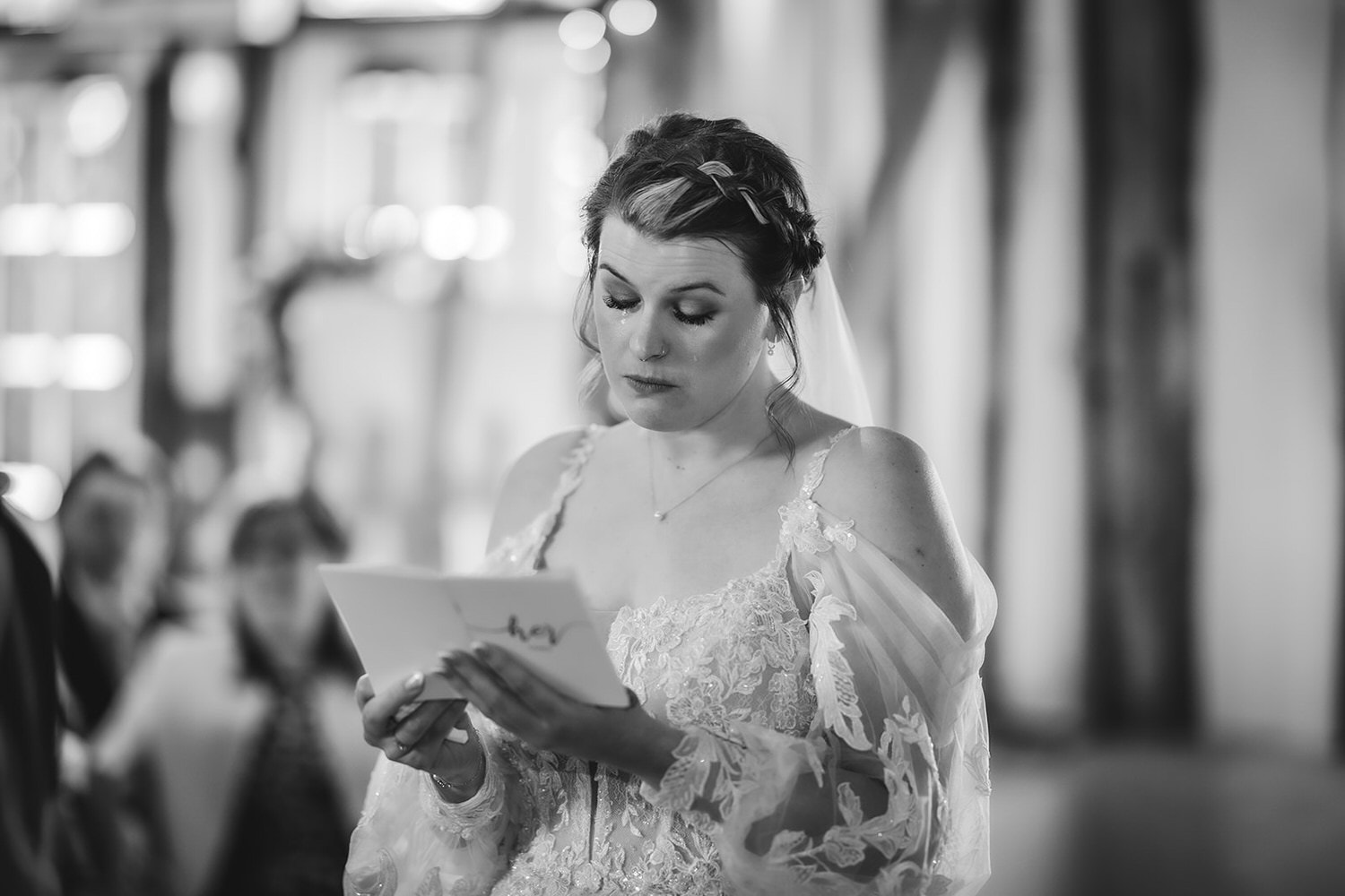 A bride in a lace dress and veil reads from a card during a wedding ceremony, looking focused and emotional. The background is softly blurred, highlighting her expression.