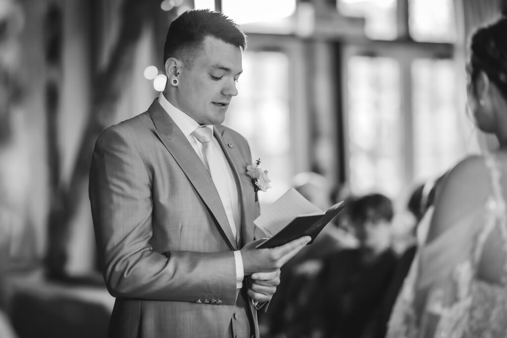 A man in a light suit reads from a notebook during a wedding ceremony, with blurred guests seated in the background and a bride partially visible in the foreground. The image is black and white.